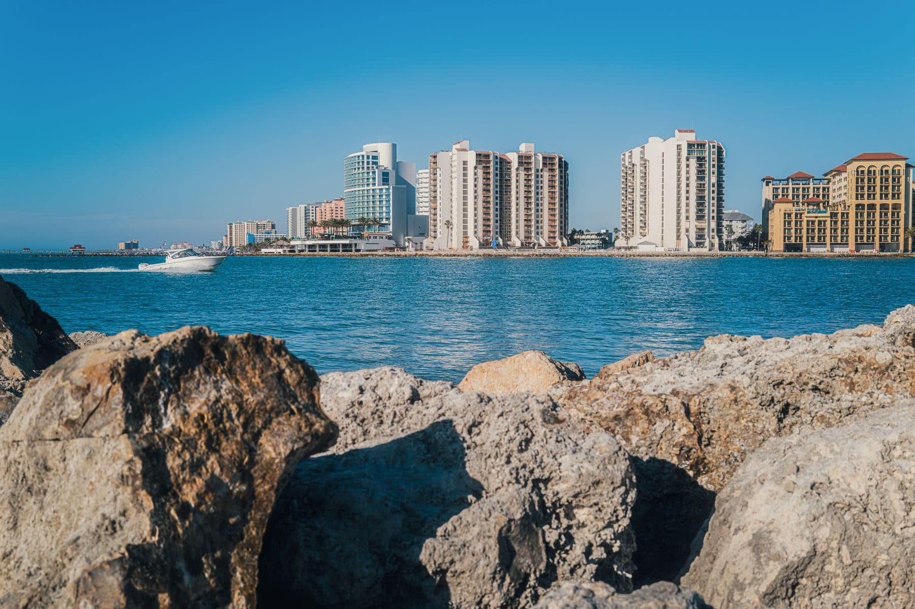 A row of high-rise condominium towers along the water, seen across a rocky shore.