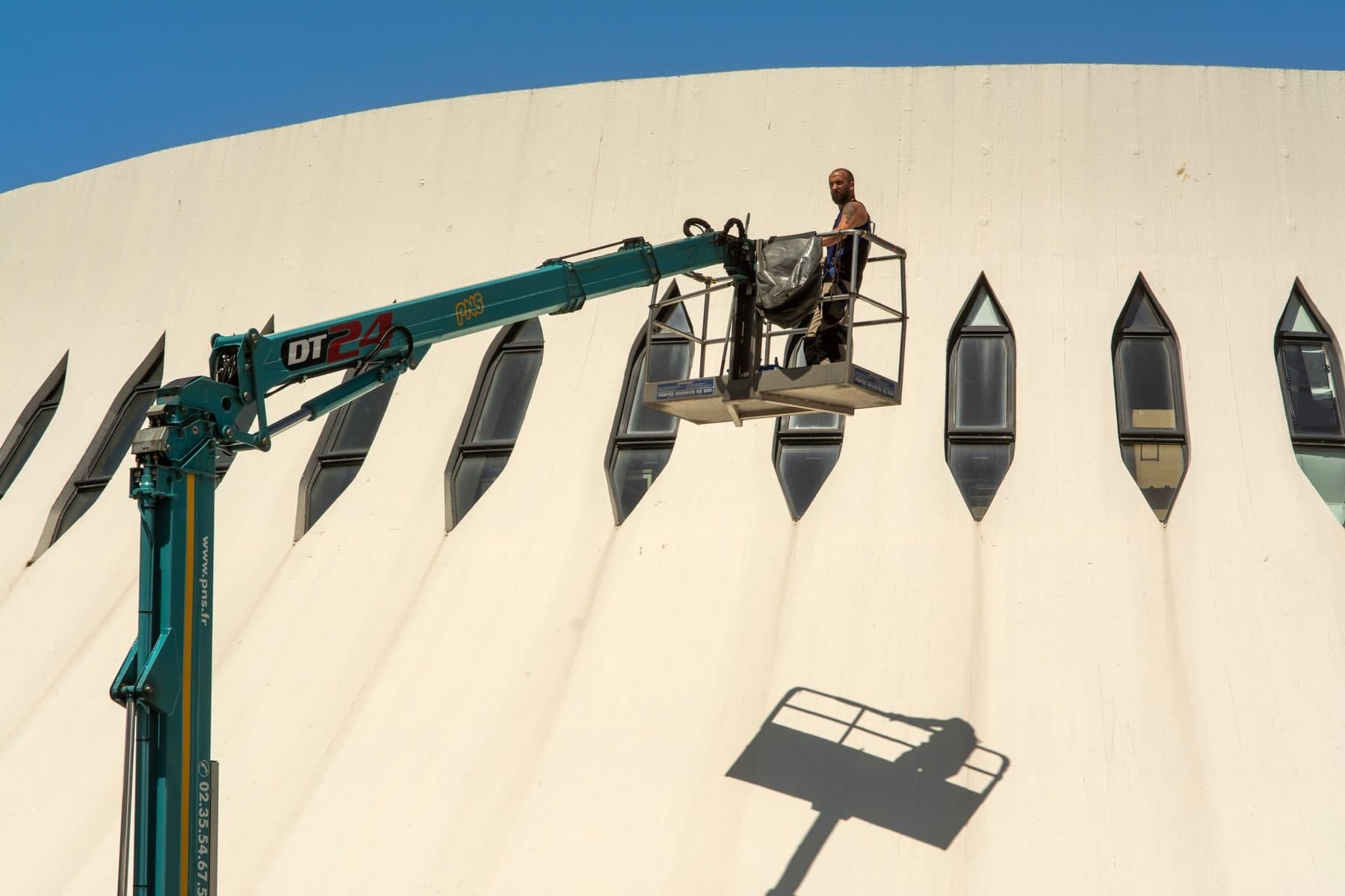 A worker in the basket of an aerial lift beside a curved cream concrete wall.