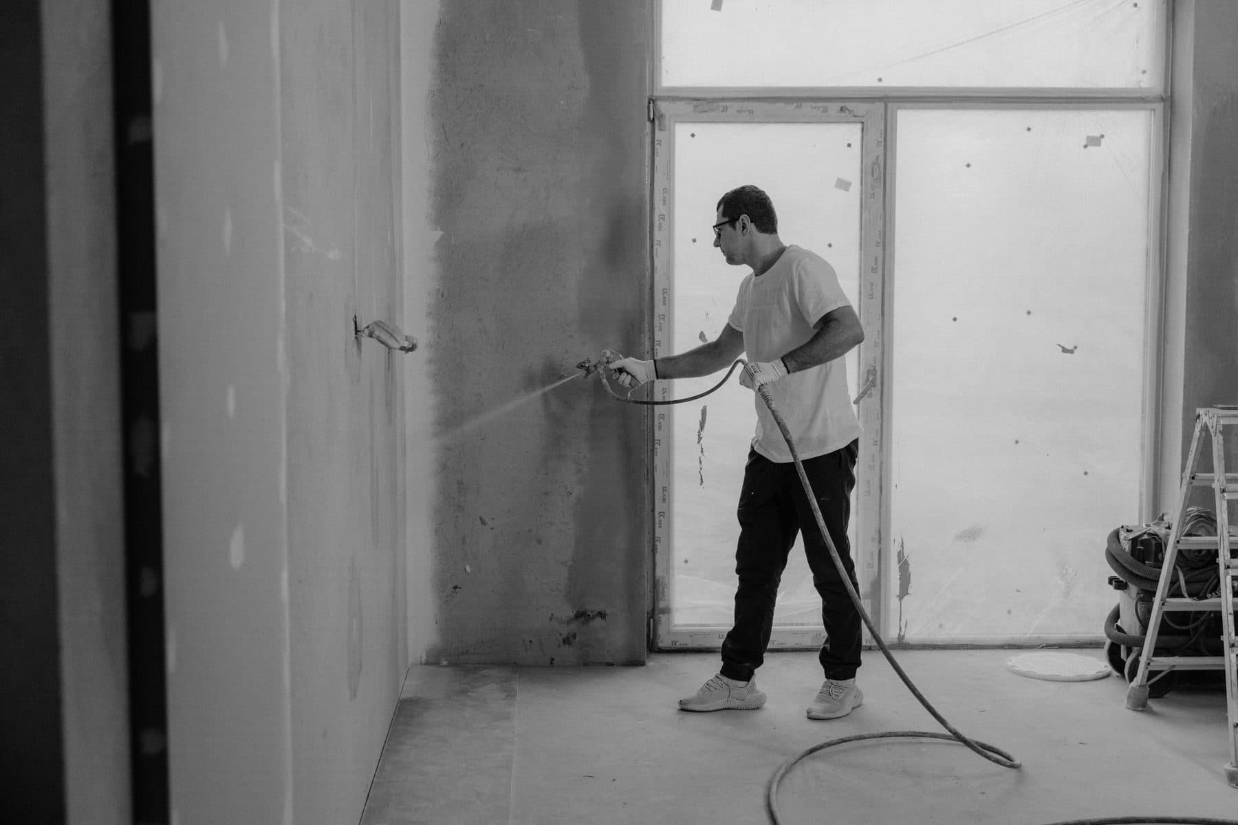 A worker spraying paint onto an interior wall with an airless spray gun, in black and white.